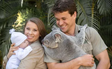 A family holding a baby and a koala, smiling in front of lush green foliage.