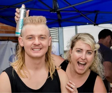 Two people smiling at a mullet competition, one holding hair spray. The man has a blonde mullet under a blue canopy.