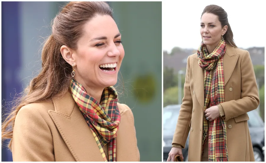 Woman in a camel coat with plaid scarf, smiling and walking outdoors.