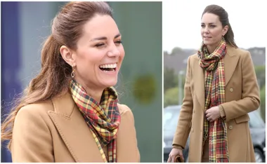 Woman in a camel coat with plaid scarf, smiling and walking outdoors.
