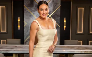A woman in a beige dress stands confidently in a stylish room with a marble table and patterned wall.