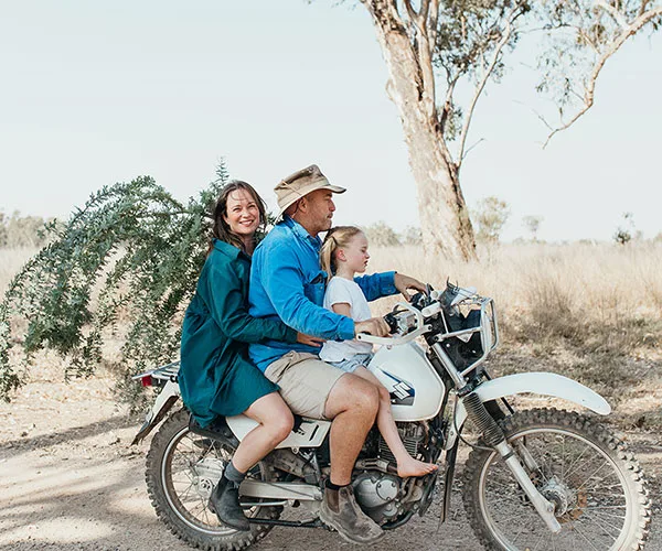 Family riding a motorcycle in a rural area, carrying a tree branch, smiling and enjoying the countryside atmosphere.
