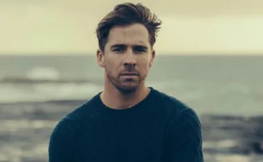 Man in a blue sweater stands by the sea with a contemplative expression.