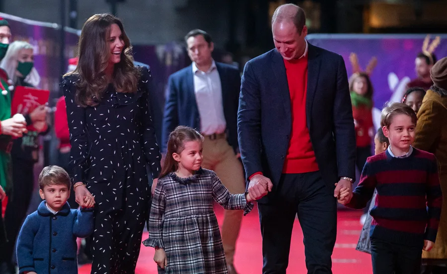 Kate and William with their children on a red carpet at a festive event, surrounded by other people.