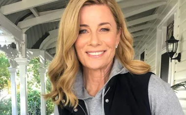 A woman with blonde hair smiling, standing on a white veranda with greenery in the background.