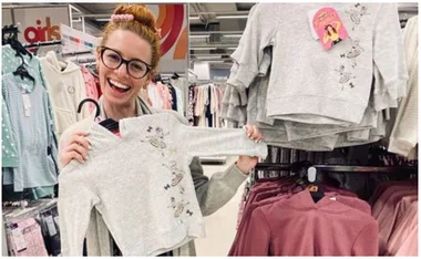 Woman smiling and holding a gray hoodie with Emma Wiggle design in a clothing store, surrounded by similar hoodies.