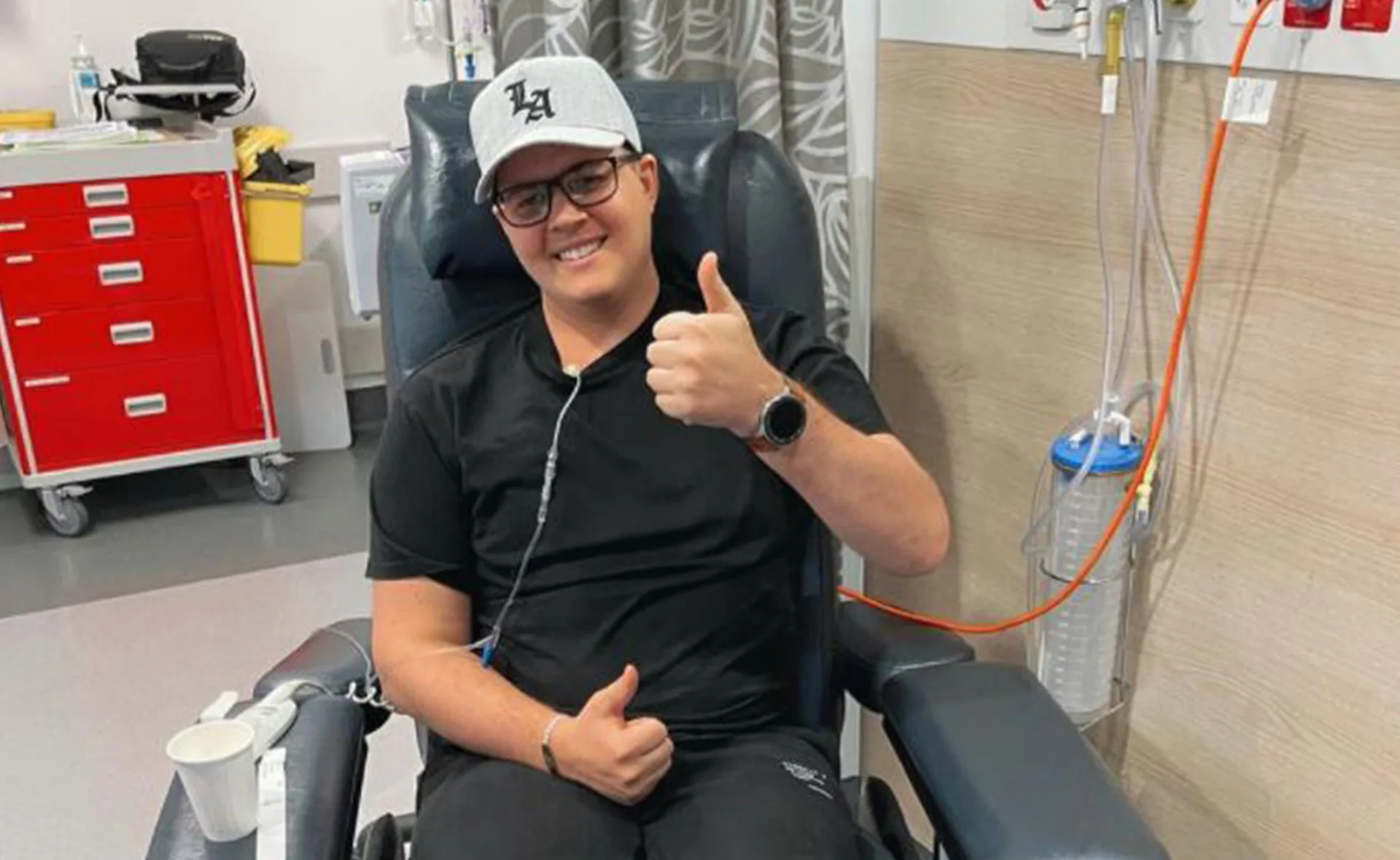 Man in a hospital chair giving a thumbs-up, wearing glasses and a cap, attached to medical equipment.