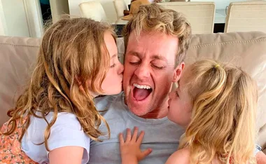 Father on couch with eyes closed, smiling as two young children kiss his cheeks.