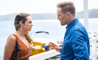 Man and woman having a serious conversation by a lakeside, with a yellow boat in the background.