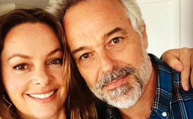 Two people smiling closely together, one with long hair, the other with gray hair and a beard.