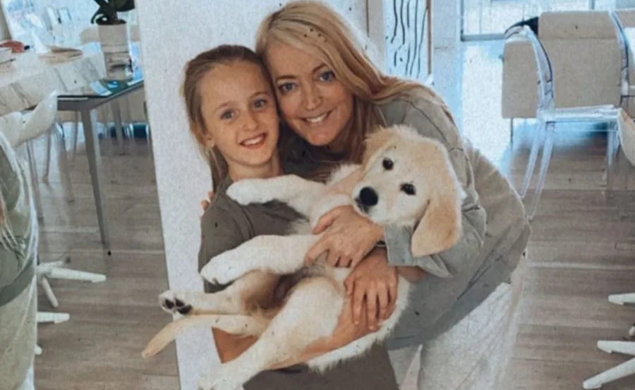 A woman and a young girl smiling while holding a fluffy puppy inside a bright, modern room.