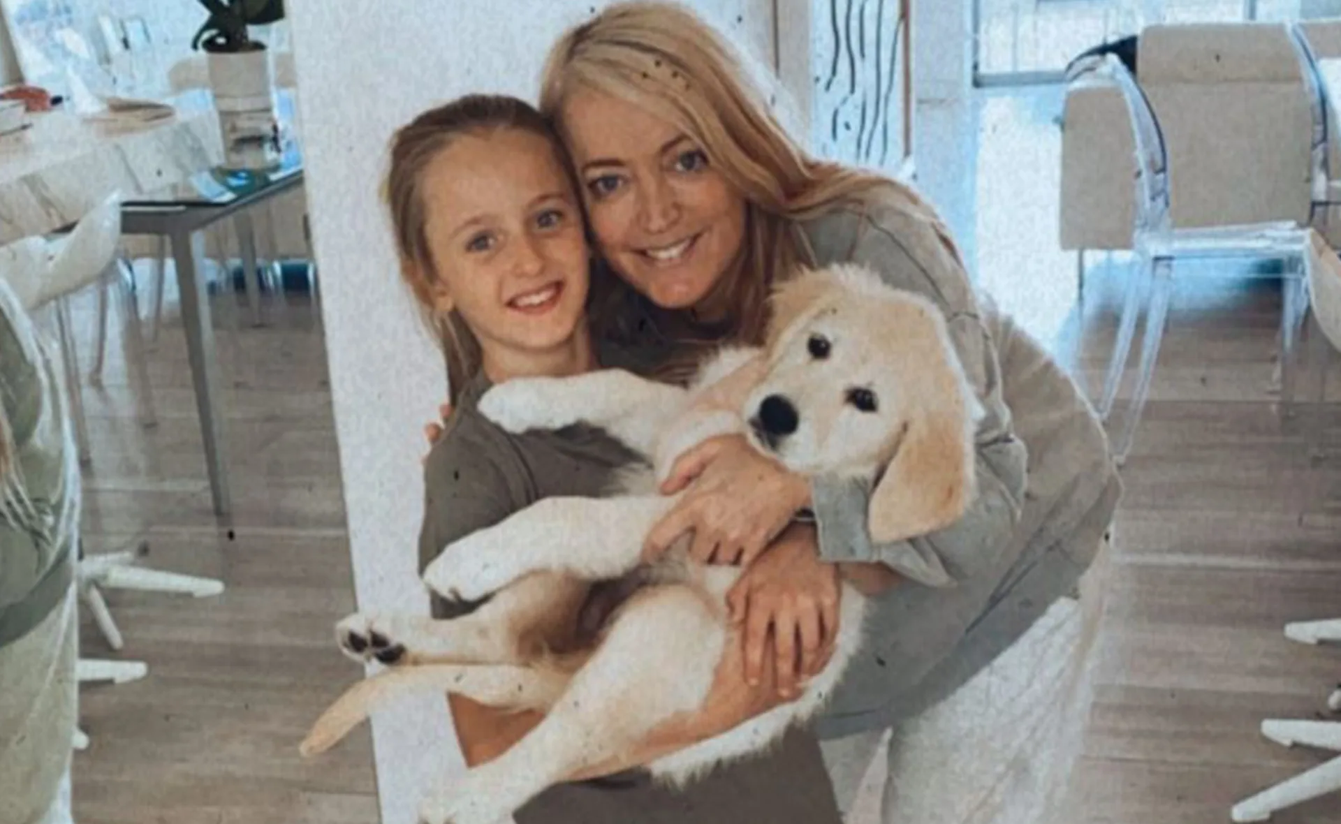 A woman and a young girl smiling while holding a fluffy puppy inside a bright, modern room.