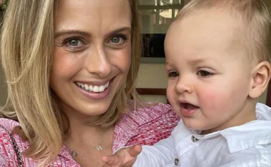 A woman in a pink outfit smiles while holding a baby in a white shirt.