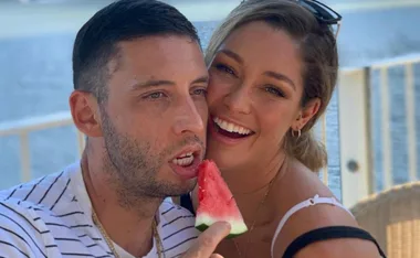 A couple smiling on a balcony, with the man holding a slice of watermelon near his mouth.