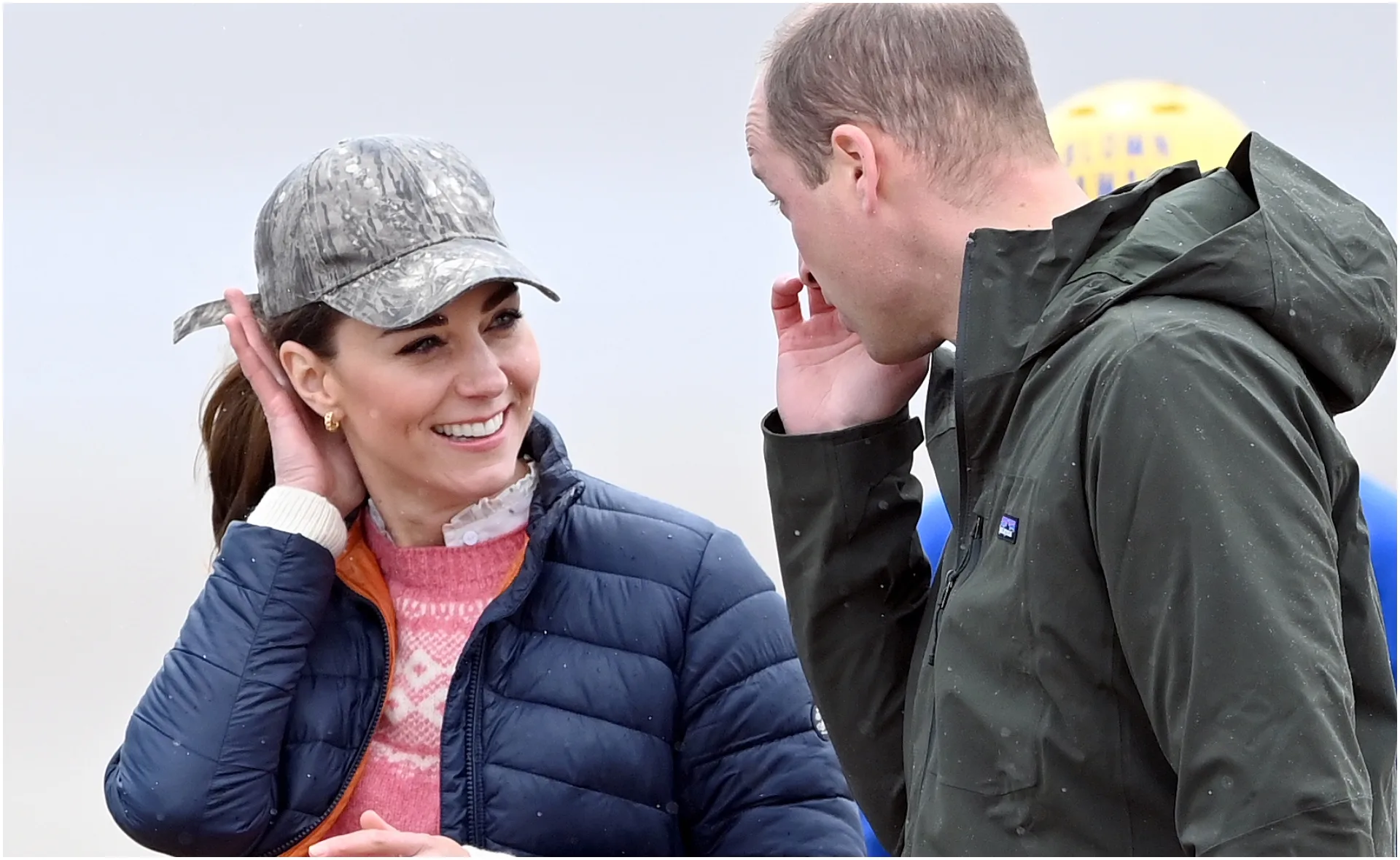 Two people outdoors, smiling and talking; one in a cap, the other in a hooded jacket, under a cloudy sky.