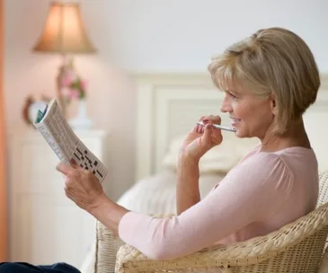A woman sitting in a chair, thoughtfully holding a pen and doing a puzzle from a magazine in a cozy room.