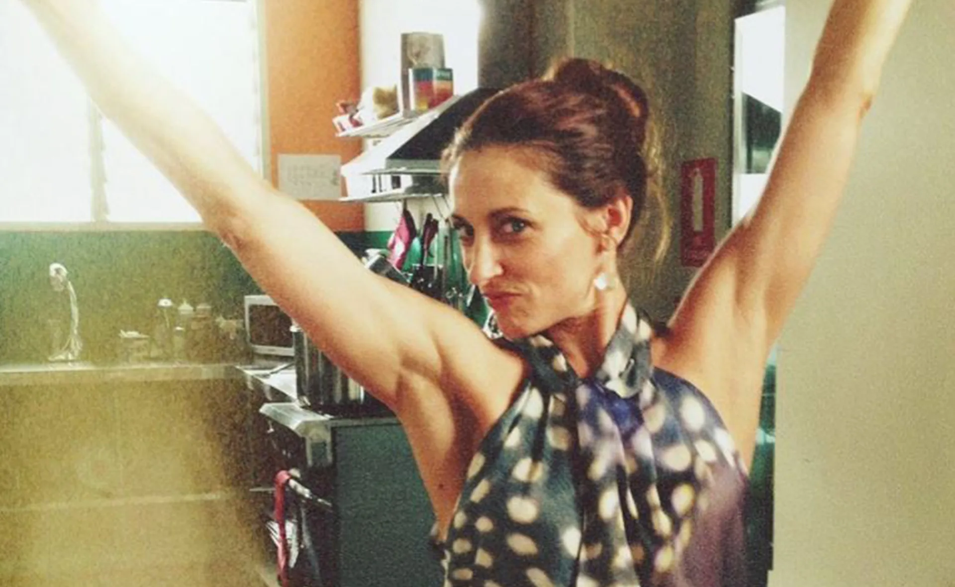 A woman with arms raised, wearing a patterned top, smiling confidently in a kitchen setting.