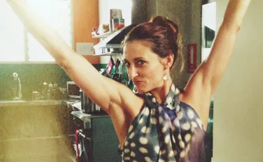 A woman with arms raised, wearing a patterned top, smiling confidently in a kitchen setting.