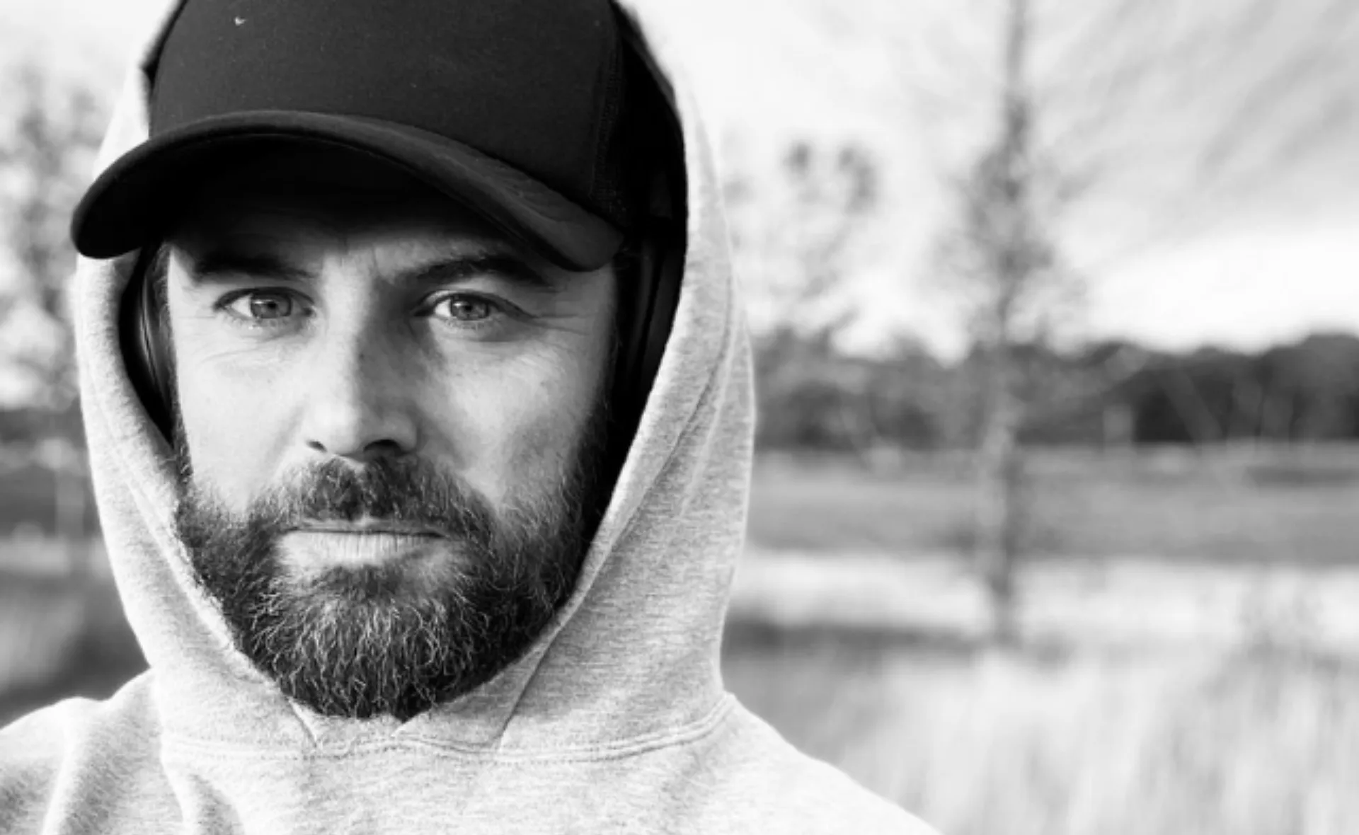 Black and white photo of a bearded man wearing a hoodie and cap outdoors.