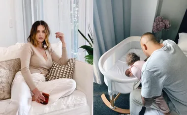 A woman sits on a couch holding a drink, while a man tends to a baby in a crib in a cozy room.
