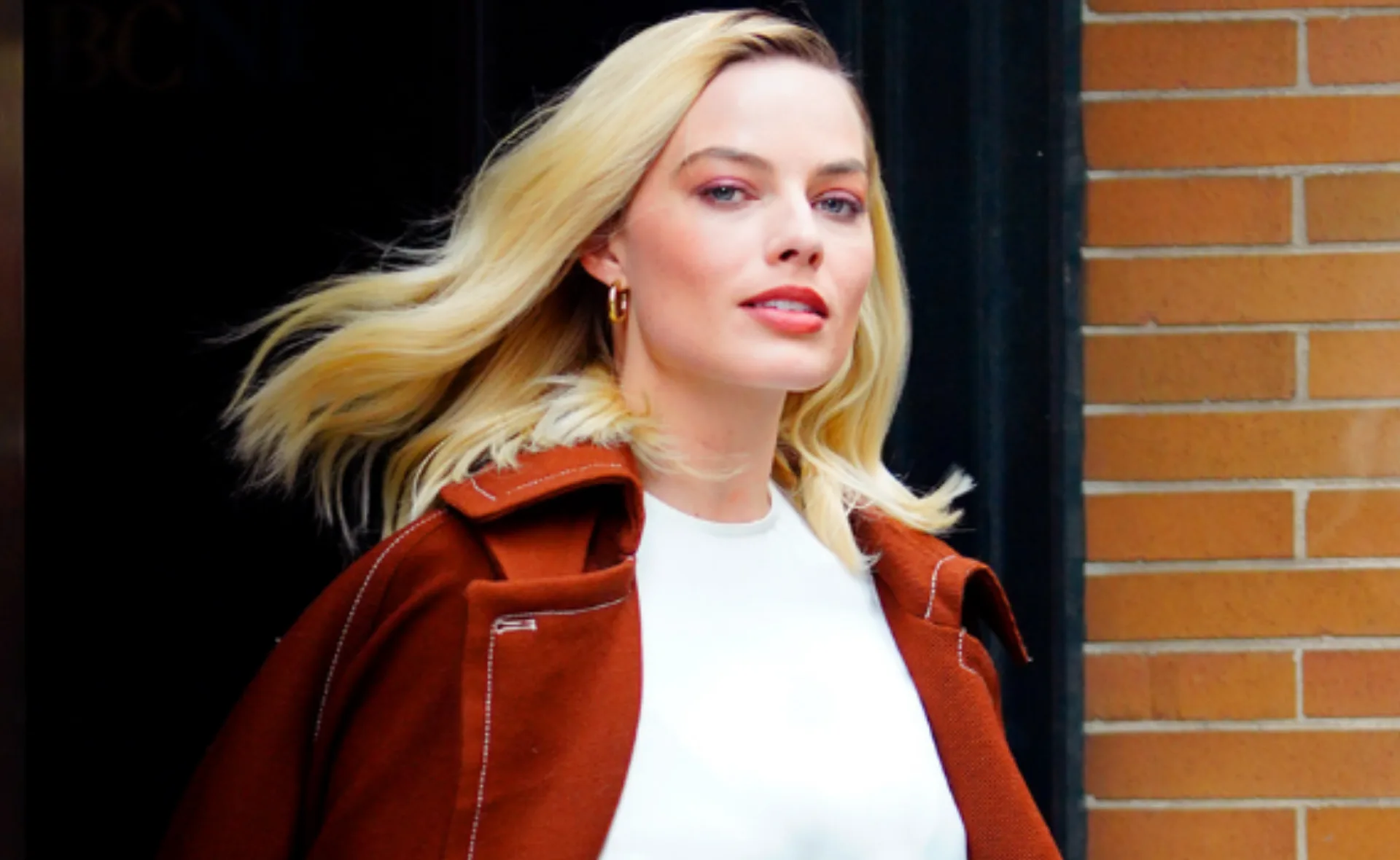 A woman with blonde hair wearing a white top and brown coat, standing against a brick wall background.