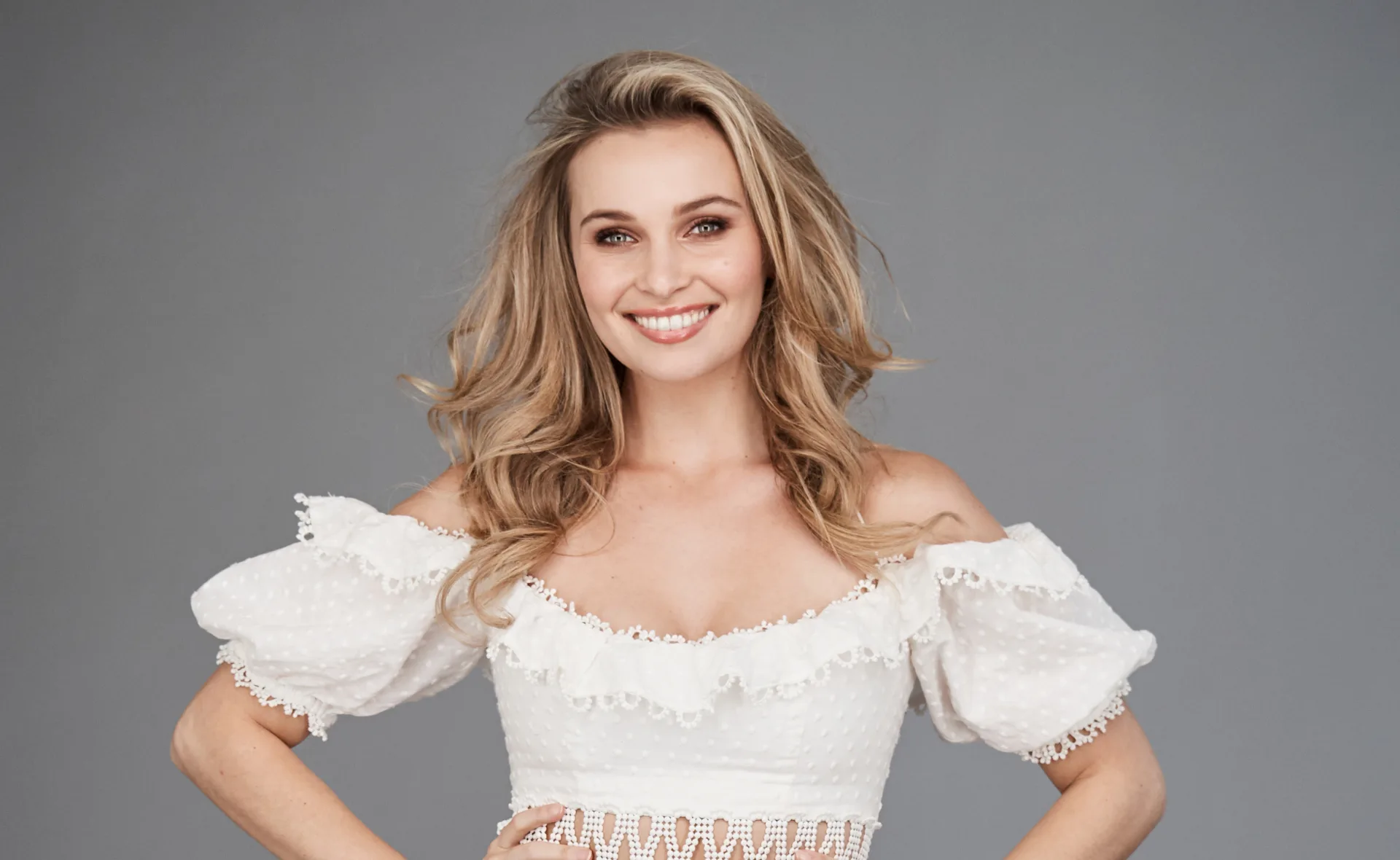Young woman smiling, wearing an off-shoulder white blouse, standing against a gray background.