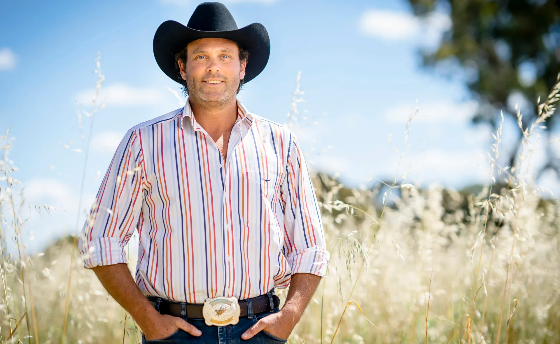 Man in striped shirt and black cowboy hat standing in a sunny field with tall grasses and a blue sky background.