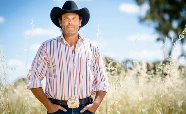 Man in striped shirt and black cowboy hat standing in a sunny field with tall grasses and a blue sky background.