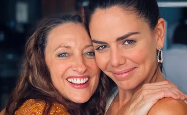 Two women smiling closely together, one with long curly hair and the other with dark hair tied back, both looking happy.