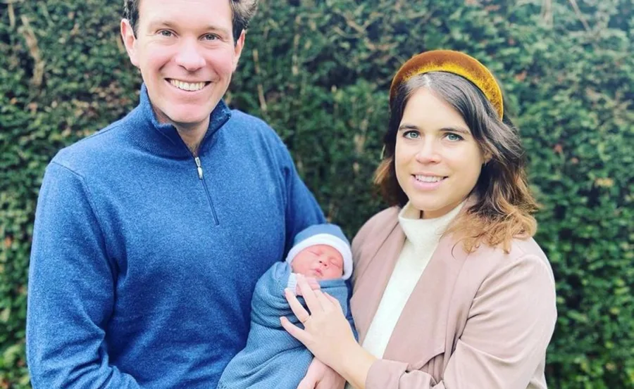 A smiling couple holds a swaddled baby outdoors, with greenery in the background.