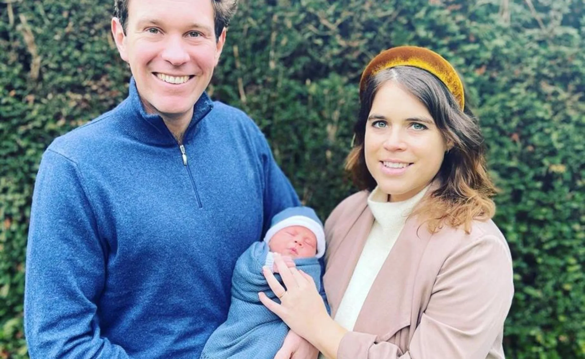 A smiling couple holds a swaddled baby outdoors, with greenery in the background.