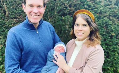 A smiling couple holds a swaddled baby outdoors, with greenery in the background.