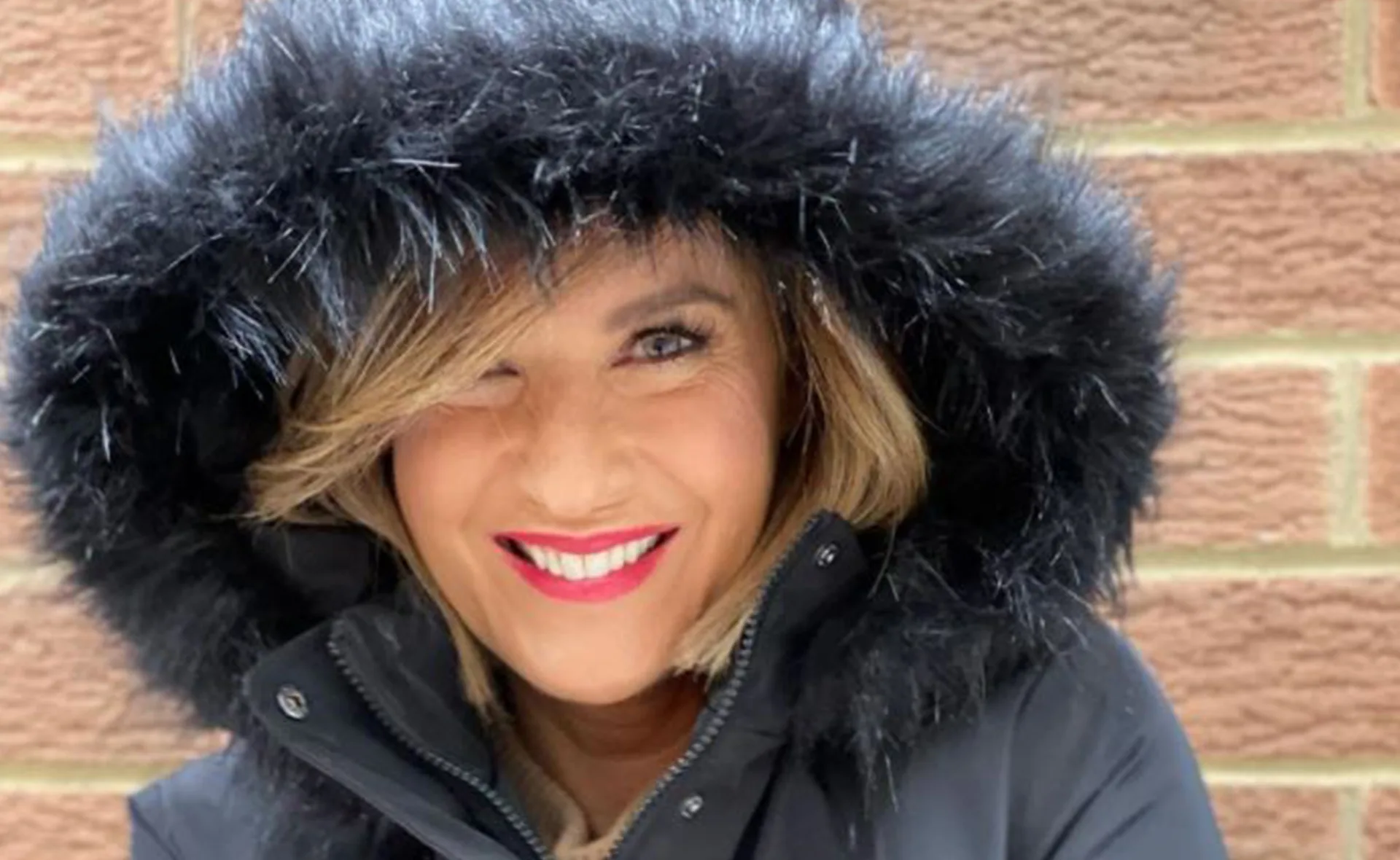 Person in a black fur-trimmed coat hood, smiling brightly against a brick wall.
