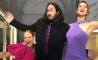 Three people joyfully posing together; one man in the middle with long hair and women in vibrant dresses on either side.