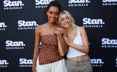Two women smiling in front of a Stan Originals backdrop, one in patterned top, the other in white tank.