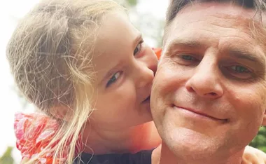 Father and daughter smiling warmly outdoors, with the daughter playfully resting her chin on his shoulder.