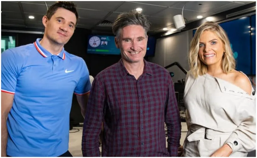 Three radio hosts smiling in a studio with microphones and screens around them.