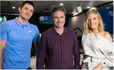 Three radio hosts smiling in a studio with microphones and screens around them.