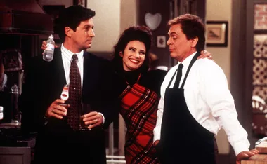Three people in a kitchen: a man in a suit holding a drink, a woman in plaid smiling, and a man in an apron.