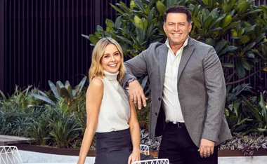 Two people smiling outdoors, with greenery in the background, both dressed in business casual attire.
