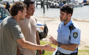 Police officer in uniform speaks to two men near a beach with a police vehicle in the background.