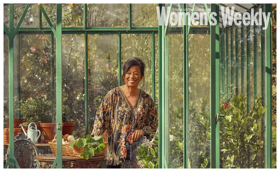 A woman smiling in a floral blouse, standing in a greenhouse surrounded by plants and potted flowers.