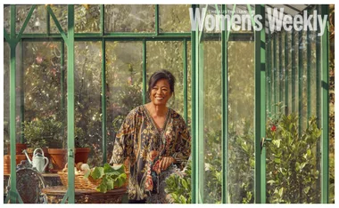 A woman smiling in a floral blouse, standing in a greenhouse surrounded by plants and potted flowers.