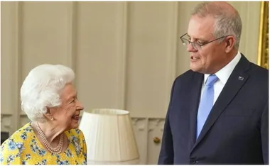 Queen Elizabeth II in a yellow dress, smiling and conversing with a suited man indoors.