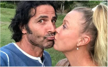 A woman and man kissing outdoors, with greenery in the background.