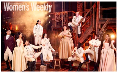 Cast of "Hamilton" Australia in period costumes, posing energetically on stage, set with ropes and wooden beams.