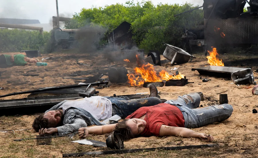 Two people lying unconscious on sandy ground amid debris and small fires.
