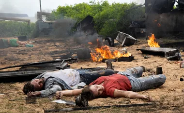 Two people lying unconscious on sandy ground amid debris and small fires.