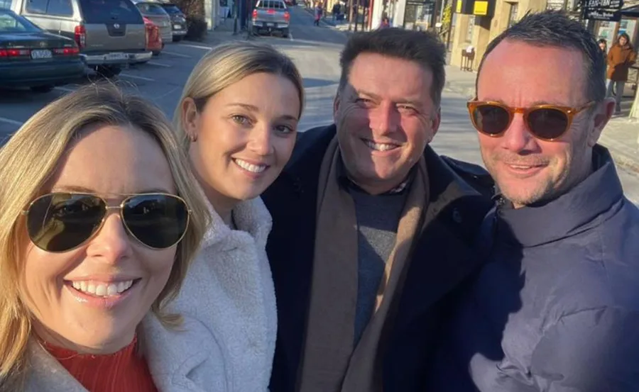 Four people smiling outdoors, wearing coats and sunglasses, in a sunny urban setting.