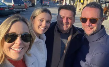 Four people smiling outdoors, wearing coats and sunglasses, in a sunny urban setting.